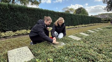 Auf dem Bild sieht man zwei junge Frauen, die gemeinsam mit einer Bürste einen Grabstein reinigen.