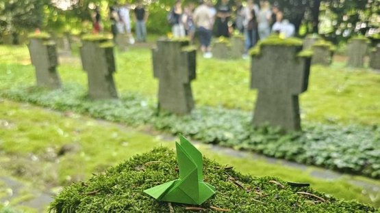Im Vordergrund ist ein Grabstein mit einer grünen Friedenstaube zu sehen. Im Hintergrund steht eine Gruppe