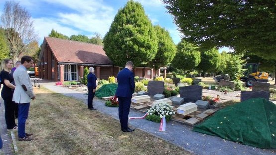 Auf dem Bild sieht man vier Holzsärge vor ausgehobenen Gräbern, dafür stehen die Vertreter des polnischen Generalkonsulats, Frau Wojda und Herr Krzeminski, mit dem gebeugtem Haupt und mit dem Rücken zum Betrachter.