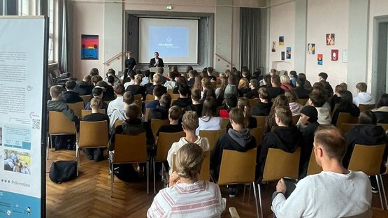 Auf dem Bild blickt man von hinten durch einen Saal über die Köpfe vieler Schülerinnen und Schüler hinweg auf eine Leinwand, vor der ein Mann einen Vortrag hält.